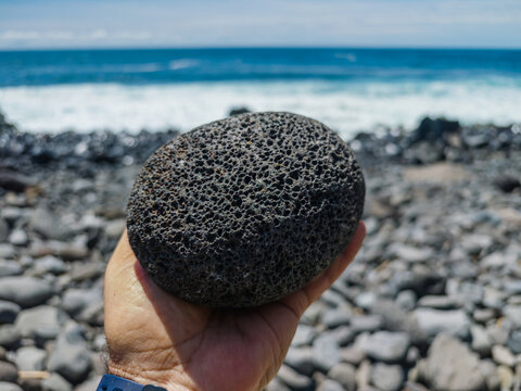 A person holding a round porous black volcanic basalt stone in their hand with a pebble beach and ocean background, Seixal, Madeira, Portugal.