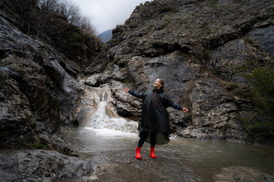 Joyful woman standing in a dramatic mountain gorge during overcast weather, wearing waterproof outdoor clothing and rubber boots. Rainy hiking, nature travel, freedom and active lifestyle concept.