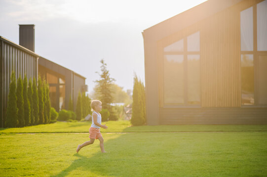 Young children running barefoot and playing together on green lawn near a modern house in warm evening sunlight. Active childhood, outdoor play, summer leisure and carefree lifestyle concept.