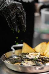 A gloved chef sprinkles chopped fresh parsley over a skillet of cooked mussels in a creamy white wine sauce, served with slices of toasted bread. © pavel siamionov