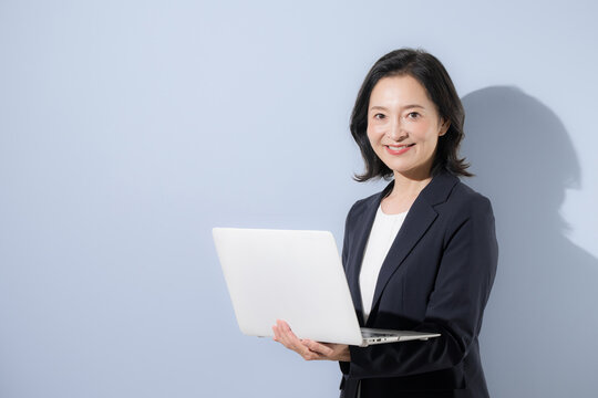 水色背景のスーツでパソコンを持つ笑顔の50代女性ビジネスイメージ、働く女性や転職やキャリアなどのカメラ目線笑顔