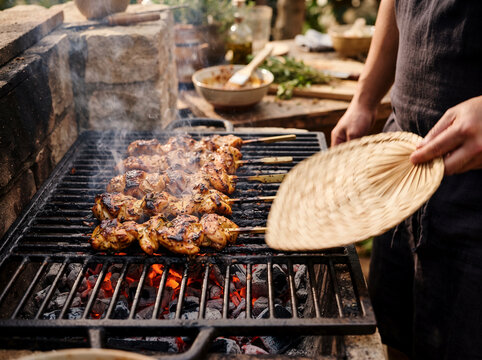 Man Grilling Chicken over Charcoal Grill with Hand Fan and Smoke Sparks