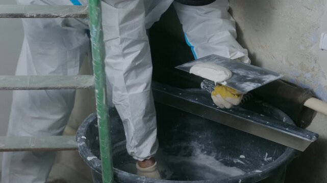 Builder taking white putty from bucket with small spatula, transferring it onto large trowel with handle for wall application. Renovation work. Worker scooping putty from bucket on trowel with spatula