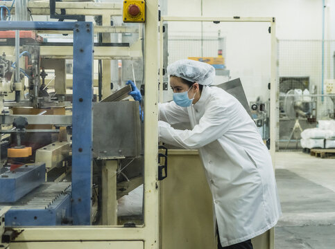 Female Factory Operator Inspecting Packaging Machine Food Process Plant Female technician in protective hygienic coat hairnet mask operating industrial packaging machinery quality assurance concept.