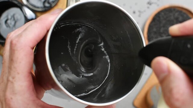 Handheld frother blends black sesame paste with milk in metal cup, creating smooth creamy base for iced cafe beverage.