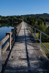 Obraz premium Narrow concrete pedestrian bridge pathway over calm lake leading towards green hills