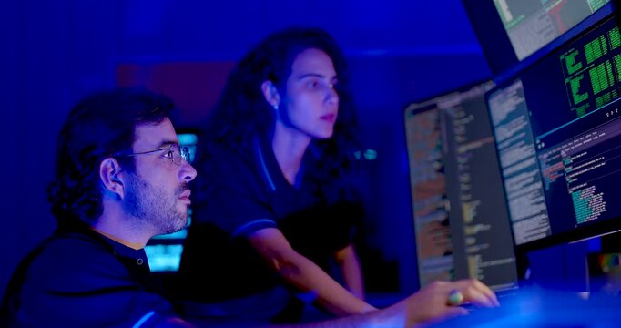 Adult male cybersecurity analyst uses mouse while female colleague studies code monitors, then points toward screen during collaborative digital system review and network security analysis at work.