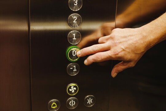 Man hand pressing a glowing ground floor zero button on a modern metal elevator control panel inside an office building or hotel close up view.