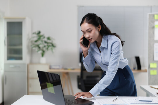 Asian businesswoman multitasking, talking on a mobile phone and typing on a laptop, standing at her office desk while managing communications and productivity