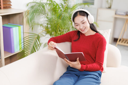 Young woman wearing white headphones, sitting comfortably on a light-colored sofa while attentively reading a book, embracing a moment of learning and leisure at home