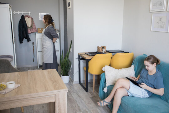 Attractive child girl using a tablet gadget while her mother does housework at home