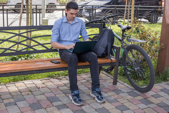A young man sits on a bench in a park using his laptop. He has a bicycle leaning against the bench and is focused on his work