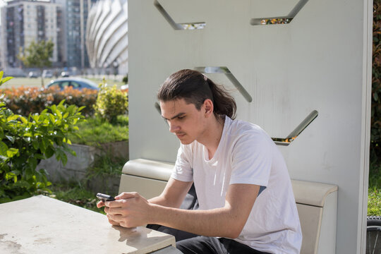 A young man in white t-shirt sits on a bench at a bus stop in an urban park. He is focused on his phone while a bicycle is parked next to him