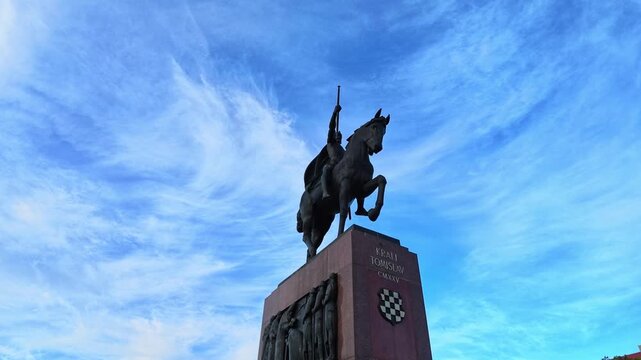 Low-angle travel video of the equestrian monument to King Tomislav in Zagreb, Croatia. The dark bronze rider and horse rise dramatically against a vivid blue sky with delicate clouds