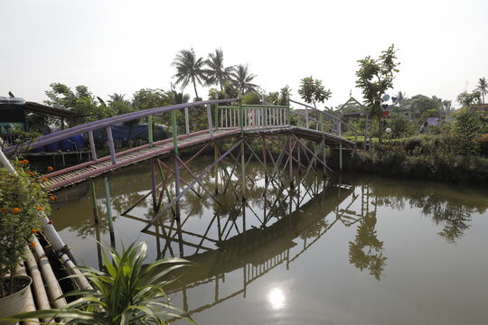 Simple handmade bamboo bridge over a small canal surrounded by green trees under a clear sky.