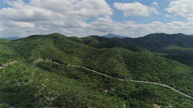 4K aerial drone view pans right over green desert mountains near La Ventana, revealing a winding road as clouds continue moving above.