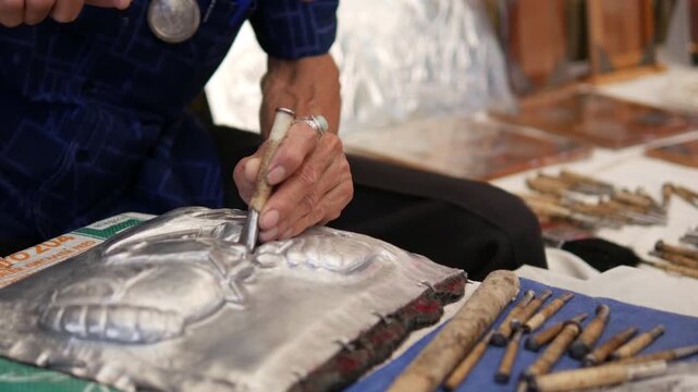 The artisan's hands are embossing a silver sheet using a hammer and punch to chase patterns.