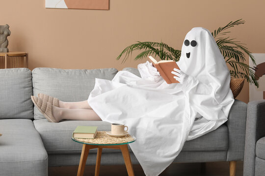 Person in a ghost costume reading a book on a gray sofa in a cozy living room, halloween and relaxation concept, a small side table with a coffee cup and another book is visible next to the sofa