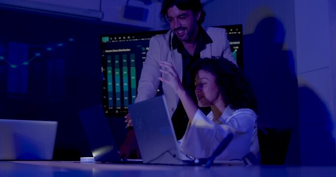 Adult female professional smiles and gestures toward laptop while male manager points at screen, exchanging ideas about performance metrics during collaborative business intelligence analysis.