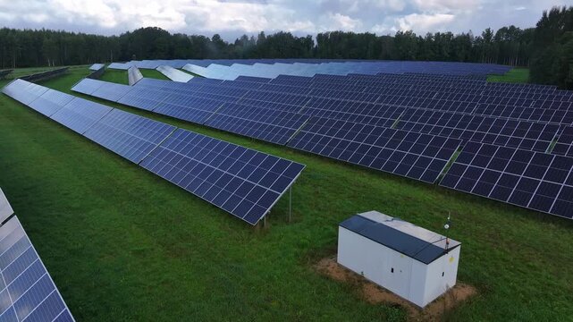 Cinematic aerial drone footage flying over an extensive solar farm with photovoltaic panels and electrical infrastructure. A technical equipment building stands among the solar arrays surrounded by gr