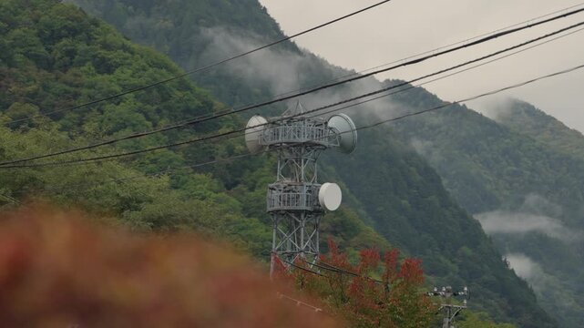 Foggy landscape featuring a telecommunication tower with satellite dishes amidst lush green mountains and power lines, with autumn colored trees providing a soft foreground in rural countryside