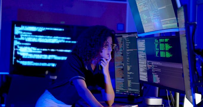 Adult female technology analyst types, pauses with hand near face, gestures toward monitor, and resumes reviewing code during cybersecurity monitoring and secure digital data analysis at work.