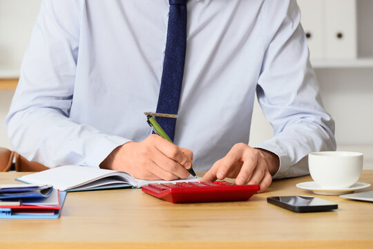 Financial planning and budget management, businessman calculating expenses and writing notes in a notebook at an office desk with a red calculator