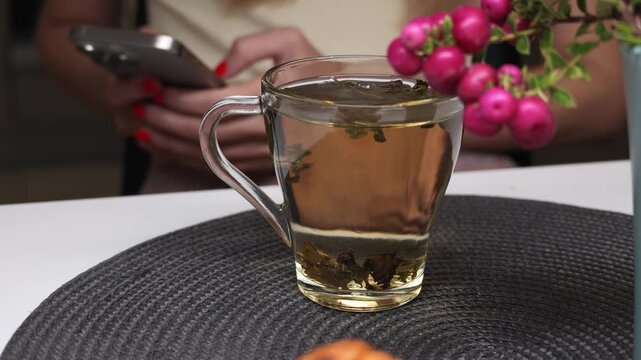 Lifestyle video showing a glass cup of herbal tea placed on a table while a woman uses a smartphone in the background