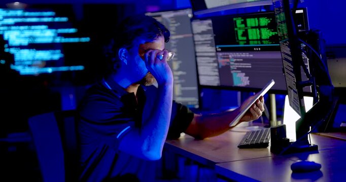 Adult male cybersecurity analyst checks tablet, adjusts glasses, then points toward code monitor while reviewing network security information and digital system data during secure operations.