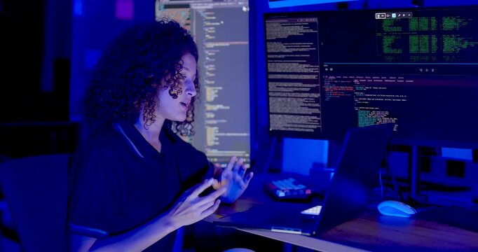 Adult female technology professional points toward source code on large monitors, discusses system findings and gestures while reviewing cybersecurity data in modern digital operations workplace.