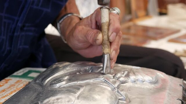 The artisan's hands are embossing a silver sheet using a hammer and punch to chase patterns.