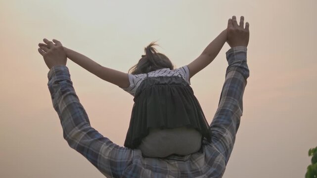 Tender moment between daughter and father, little girl riding high on her father's shoulders with trust and joy in warm golden sunset light.
