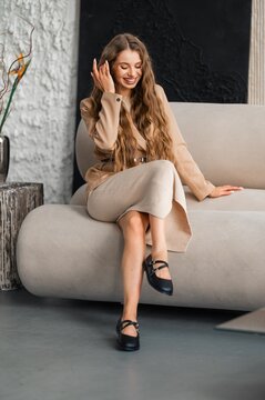 Beige blazer dress woman seated on plush sofa smiling indoors with long wavy hair, relaxed fashion lifestyle portrait