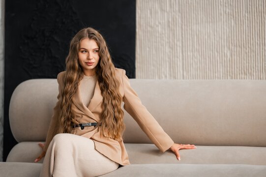 Portrait of woman in beige blazer and matching knit dress seated on light gray sofa in modern two tone interior with textured beige and black wall soft natural light