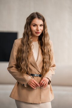 Portrait of woman with long wavy brunette hair wearing beige blazer and belt, poised in neutral studio setting with soft natural lighting, calm confident expression, elegant fashion portrait