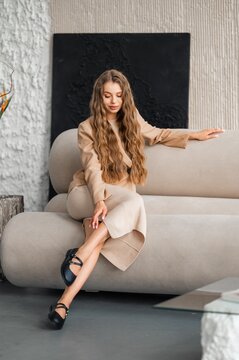 Elegant woman with long wavy hair seated on beige sofa in contemporary living room featuring textured wall, soft warm lighting, calm relaxed pose, neutrals, stylish outfit, cozy home interior decor