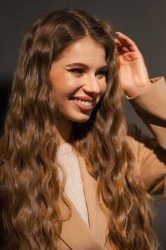 Smiling woman with long wavy brunette hair wearing beige coat photographed in warm natural light, closeup portrait emphasizing cheerful expression, skin tone, and soft textured background