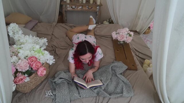 Cozy Morning: Young Woman Reading a Book in Bed Surrounded by Flowers and Candles