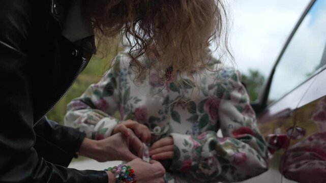 Mother daughter bonding, family travel, mom helping young girl button floral coat next to car outdoors