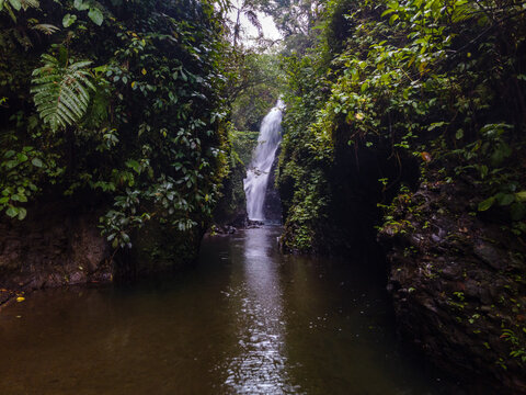 Aerial View of Hidden Lesung Waterfall Deep in the Jungle on the Slopes of Mount Slamet, A Cave-Like Tunnel Formed by Wild Trees, Moss, and Rocks Frames the Waterfall in Indonesia.