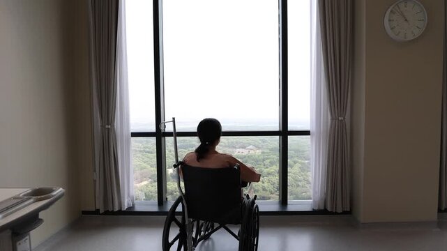 An Asian female patient sitting alone in a wheelchair inside a spacious hospital room with an IV pole attached, looking out the big window overlooking a forest view.