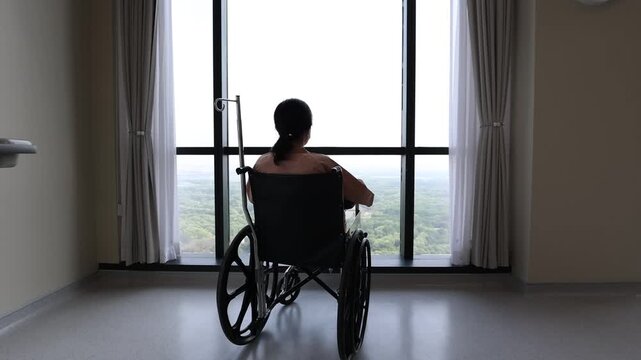 An Asian female patient sitting alone in a wheelchair inside a spacious hospital room with an IV pole attached, looking out the big window overlooking a forest view.