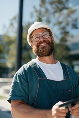 Fototapeta premium Smiling bearded construction worker wearing a white helmet and overalls outdoors for builder profile and industrial portrait banner