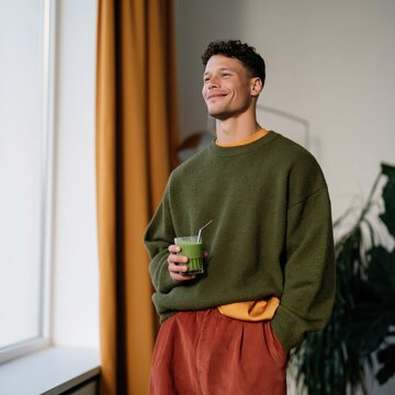 Casual man with green smoothie by window in warm interior for lifestyle poster and wellness banner design