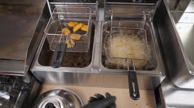 Close-up of French fries and nuggets being cooked in a deep fryer filled with boiling oil. In the kitchen, potato chips are being fried in a steel pan, along with nuggets.