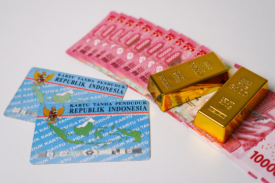 Two Indonesian national identity cards, KTP. next to fine gold bars and a fan of 100000 Rupiah banknotes on a white background.