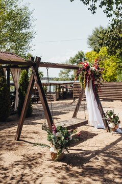 Photo of a wooden swing on a sand beach in a park with baskets of fresh colorful flowers on a wooden background.