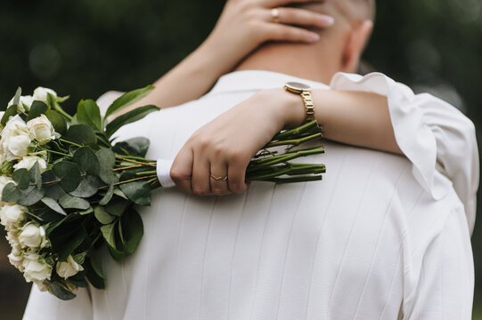 Wedding photo of a bride holding a bouquet of roses and hugging her groom from behind outdoors.