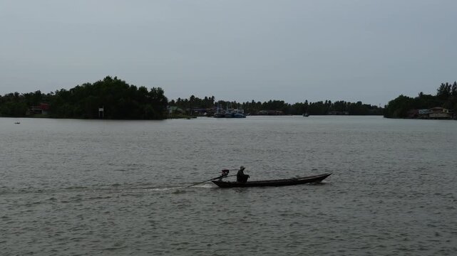 A local resident navigates a traditional motorized long-tail boat along the Tapi River during a cloudy summer day in Surat Thani, Thailand
