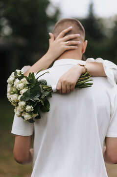 Photo of a bride holding a bouquet of roses and hugging her groom from behind outdoors.